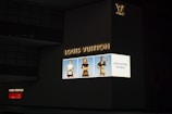 A large, illuminated advertisement for Louis Vuitton is displayed on the side of a dark building. The ad features three models showcasing handbags, dressed in stylish clothing. The Louis Vuitton logo is prominently featured, along with text in both English and another language.