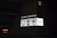 A large, illuminated advertisement for Louis Vuitton is displayed on the side of a dark building. The ad features three models showcasing handbags, dressed in stylish clothing. The Louis Vuitton logo is prominently featured, along with text in both English and another language.