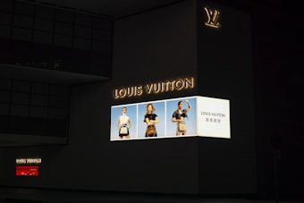 A large, illuminated advertisement for Louis Vuitton is displayed on the side of a dark building. The ad features three models showcasing handbags, dressed in stylish clothing. The Louis Vuitton logo is prominently featured, along with text in both English and another language.