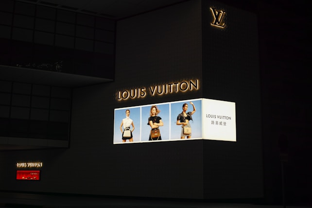 A large, illuminated advertisement for Louis Vuitton is displayed on the side of a dark building. The ad features three models showcasing handbags, dressed in stylish clothing. The Louis Vuitton logo is prominently featured, along with text in both English and another language.