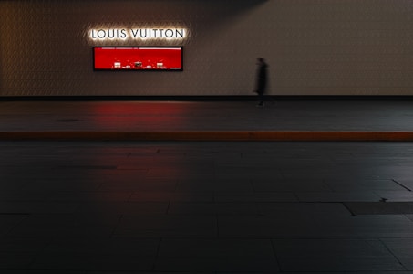 A blurred figure walks past the illuminated Louis Vuitton store window displaying products against a striking red backdrop. The scene is set against a textured wall with a geometric pattern.
