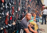 A busy market scene with a variety of leather shoes, mostly black, displayed on a wall. Several men are seated in front of the shoes, with one prominent figure in the foreground wearing a black shirt. The market stall extends into the distance with more individuals visible.