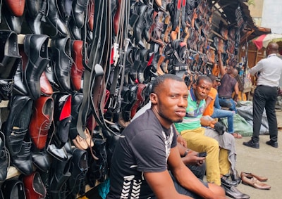 A busy market scene with a variety of leather shoes, mostly black, displayed on a wall. Several men are seated in front of the shoes, with one prominent figure in the foreground wearing a black shirt. The market stall extends into the distance with more individuals visible.