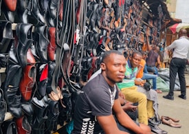A busy market scene with a variety of leather shoes, mostly black, displayed on a wall. Several men are seated in front of the shoes, with one prominent figure in the foreground wearing a black shirt. The market stall extends into the distance with more individuals visible.