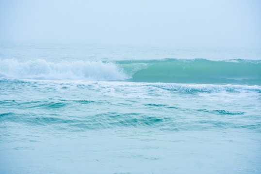 A tranquil scene of ocean waves gently rolling towards the shore. The water is a soft blend of blue and green, with subtle white foam cresting over the tops of the waves. The horizon blends seamlessly into a misty sky, creating a serene and calming atmosphere.