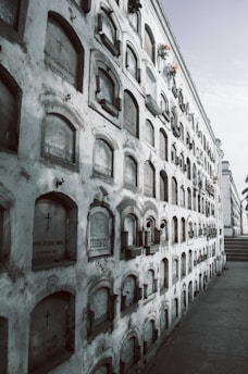 A long wall of mausoleums arranged in a grid pattern, with inscriptions and cross symbols on many of the plaques. Some mausoleums have flower arrangements attached, adding color to the otherwise gray structure. The wall extends into the distance, creating a sense of depth.
