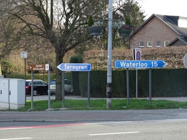 A roadside scene with multiple directional signs. Two blue signs point towards Tervuren and Waterloo, with a distance of 15 units indicated for Waterloo. A brown sign points towards a sports hall. Vehicles are parked by the roadside, and there's a lamppost with hanging flower pots. Residential houses and a hedge in the background contribute to a suburban atmosphere.