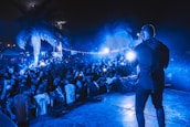 A vibrant outdoor concert scene at night with a performer on stage facing a large, energetic crowd. The atmosphere is illuminated by blue lights, and people in the audience are capturing the moment with their phones. Palm trees are visible, adding a tropical vibe to the event.