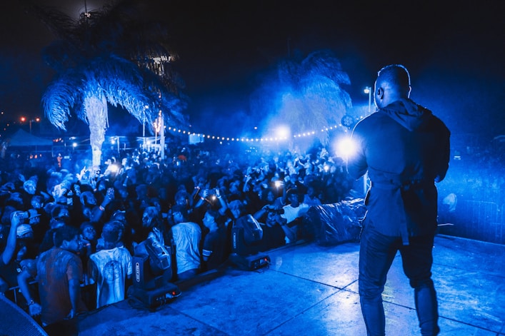 A vibrant outdoor concert scene at night with a performer on stage facing a large, energetic crowd. The atmosphere is illuminated by blue lights, and people in the audience are capturing the moment with their phones. Palm trees are visible, adding a tropical vibe to the event.