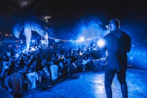 A vibrant outdoor concert scene at night with a performer on stage facing a large, energetic crowd. The atmosphere is illuminated by blue lights, and people in the audience are capturing the moment with their phones. Palm trees are visible, adding a tropical vibe to the event.