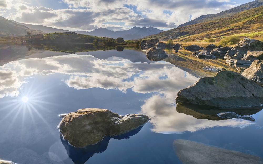 montanha marrom e verde ao lado do lago sob nuvens brancas e céu azul durante o dia