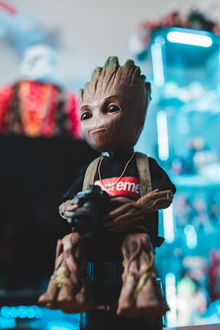 A figurine of a character with a wooden-like texture is dressed in a black shirt with a red logo. The figure appears to be holding a camera. Bright colors and lights can be seen in the background, giving a modern and vibrant look.