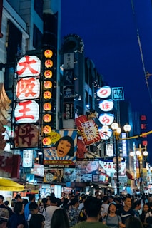 A bustling urban street filled with a crowd of people under a vibrant array of illuminated Japanese signs and billboards. The street is animated with activity, with bright neon lights advertising various shops, restaurants, and entertainment venues. Characters and bold graphics dominate the signs, contributing to the lively atmosphere.