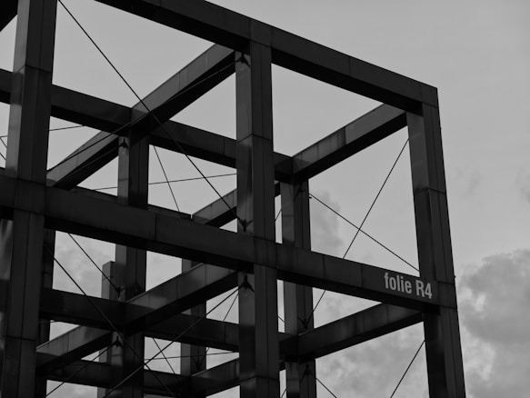 A geometric structure made of metal beams, forming a series of connected cubes. Tension wires run through and across the structure, providing additional support. The sky in the background is overcast, with some visible clouds.