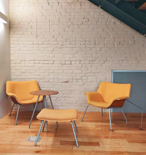 A cozy seating area featuring two orange upholstered chairs and a matching footstool arranged around a small round wooden table. The setting is indoors with a brick wall painted white and a wooden floor. The area is lit by natural or ambient light, creating a warm atmosphere.