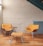 A cozy seating area featuring two orange upholstered chairs and a matching footstool arranged around a small round wooden table. The setting is indoors with a brick wall painted white and a wooden floor. The area is lit by natural or ambient light, creating a warm atmosphere.