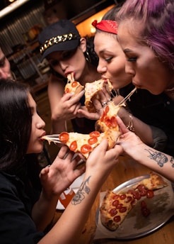 Four individuals are enjoying slices of pepperoni pizza, stretching gooey cheese between their hands. They are gathered around a wooden table, sharing a plate of pizza with vibrant toppings. The scene conveys a sense of camaraderie and fun with visible tattoos on some arms and casual attire.