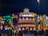 A vibrant nighttime scene features a grand, illuminated building with the word 'AMERICAS' prominently displayed on top. This structure is flanked by colorful, artistic designs that include a large decorative totem pole on the left and intricate sculptural elements on the right. The scene is lively with numerous people walking and gathering in front of the building.
