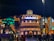 A vibrant nighttime scene features a grand, illuminated building with the word 'AMERICAS' prominently displayed on top. This structure is flanked by colorful, artistic designs that include a large decorative totem pole on the left and intricate sculptural elements on the right. The scene is lively with numerous people walking and gathering in front of the building.