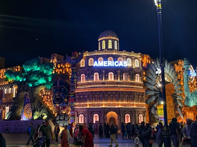 A vibrant nighttime scene features a grand, illuminated building with the word 'AMERICAS' prominently displayed on top. This structure is flanked by colorful, artistic designs that include a large decorative totem pole on the left and intricate sculptural elements on the right. The scene is lively with numerous people walking and gathering in front of the building.