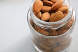 Close-up of a jar of buttr's espresso brown almond butter on a smooth stone slab, surrounded by whole almonds and a scattering of cocoa nibs in soft natural light.