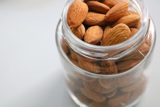 A transparent glass jar filled with whole almonds is placed on a light surface. The almonds are tightly packed, displaying a variety of rich brown tones with natural textures.