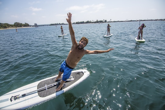 A group of people are paddleboarding on a sunny day in a large body of water. In the foreground, a man playfully leans off his paddleboard with a wide smile, while others in the distance paddle calmly. The scene conveys a sense of fun and leisure, set against a backdrop of a clear sky and distant shore.