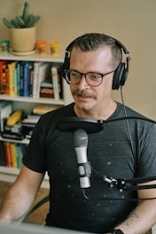 A person with glasses and headphones is sitting in front of a microphone, appearing to be engaged in a recording or podcast session. Behind them is a bookshelf filled with various books, and a cactus plant is placed on top of it. The setting is indoors, giving a cozy and professional vibe.
