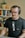 A person with glasses and headphones is sitting in front of a microphone, appearing to be engaged in a recording or podcast session. Behind them is a bookshelf filled with various books, and a cactus plant is placed on top of it. The setting is indoors, giving a cozy and professional vibe.
