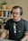 A person with glasses and headphones is sitting in front of a microphone, appearing to be engaged in a recording or podcast session. Behind them is a bookshelf filled with various books, and a cactus plant is placed on top of it. The setting is indoors, giving a cozy and professional vibe.