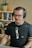 A person with glasses and headphones is sitting in front of a microphone, appearing to be engaged in a recording or podcast session. Behind them is a bookshelf filled with various books, and a cactus plant is placed on top of it. The setting is indoors, giving a cozy and professional vibe.
