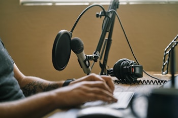 A close-up of a person typing on a keyboard, with a professional microphone and a pop filter prominently positioned in the foreground. Nearby, a pair of headphones rests on the desk. The scene suggests a podcast or audio recording setup.