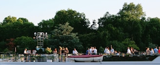A group of people gathered on a boat deck, enjoying a social event against a backdrop of lush green trees. The setting appears relaxed and pleasant, with guests conversing and taking in the natural surroundings. The boat has a sleek design, with visible guardrails and a small rescue boat on top.