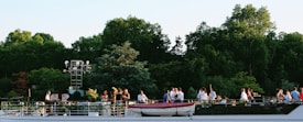 A group of people gathered on a boat deck, enjoying a social event against a backdrop of lush green trees. The setting appears relaxed and pleasant, with guests conversing and taking in the natural surroundings. The boat has a sleek design, with visible guardrails and a small rescue boat on top.