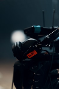 A close-up view of professional audio-visual equipment, including a pair of headphones placed on what appears to be a camera or related device. The equipment has a note attached with the name 'ANDY'. The background is dark and blurred.