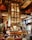 A lavish bar area with art deco architectural elements featuring intricate patterns and ornate designs. The central focus is a large, elegant bar counter surrounded by bar stools. The back of the bar showcases an impressive, illuminated wine rack reaching toward the high ceiling. Rich wood tones and golden accents dominate the decor, creating a luxurious ambiance. Tall windows allow natural light to enter, enhancing the opulence of the space.