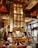 A lavish bar area with art deco architectural elements featuring intricate patterns and ornate designs. The central focus is a large, elegant bar counter surrounded by bar stools. The back of the bar showcases an impressive, illuminated wine rack reaching toward the high ceiling. Rich wood tones and golden accents dominate the decor, creating a luxurious ambiance. Tall windows allow natural light to enter, enhancing the opulence of the space.