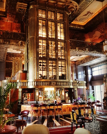 A lavish bar area with art deco architectural elements featuring intricate patterns and ornate designs. The central focus is a large, elegant bar counter surrounded by bar stools. The back of the bar showcases an impressive, illuminated wine rack reaching toward the high ceiling. Rich wood tones and golden accents dominate the decor, creating a luxurious ambiance. Tall windows allow natural light to enter, enhancing the opulence of the space.