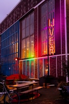 A colorful building facade with large glass windows and a neon sign that reads 'IJVER' in bright red letters. The structure has a mix of exposed brick and vibrant lighting illuminating it in shades of blue, pink, and orange. In front of the building are outdoor seating arrangements with wooden benches and tables, along with decorative elements such as potted plants and a bicycle.