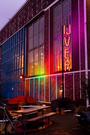 A colorful building facade with large glass windows and a neon sign that reads 'IJVER' in bright red letters. The structure has a mix of exposed brick and vibrant lighting illuminating it in shades of blue, pink, and orange. In front of the building are outdoor seating arrangements with wooden benches and tables, along with decorative elements such as potted plants and a bicycle.