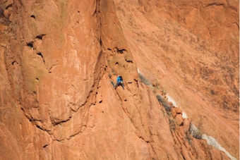 A person dressed in blue clothing is climbing a large reddish-brown rock face, likely a sandstone cliff. The setting is rugged and remote, suggesting an outdoor adventure or extreme sport.