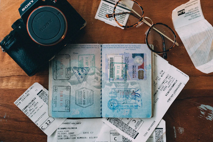 An arrangement of travel-related items including a Sony camera, an open passport with numerous stamps, flight tickets, and a pair of eyeglasses on a wooden surface.