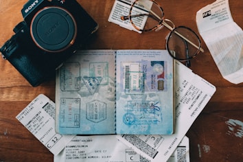 An arrangement of travel-related items including a Sony camera, an open passport with numerous stamps, flight tickets, and a pair of eyeglasses on a wooden surface.