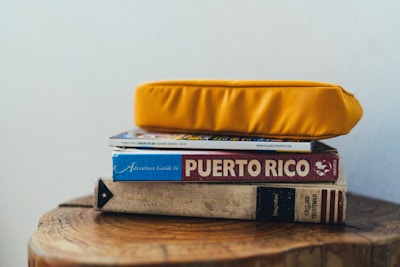A stack of books and a magazine is topped with a bright yellow cushion. The titles visible include a guide to Puerto Rico and an adventure guidebook. The books are placed on a wooden surface with natural wood grain visible.