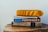 A stack of books and a magazine is topped with a bright yellow cushion. The titles visible include a guide to Puerto Rico and an adventure guidebook. The books are placed on a wooden surface with natural wood grain visible.