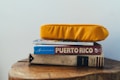 A stack of books and a magazine is topped with a bright yellow cushion. The titles visible include a guide to Puerto Rico and an adventure guidebook. The books are placed on a wooden surface with natural wood grain visible.