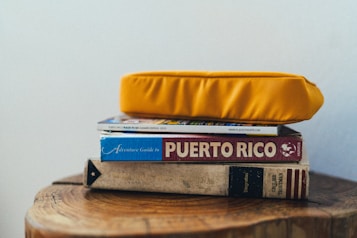 A stack of books and a magazine is topped with a bright yellow cushion. The titles visible include a guide to Puerto Rico and an adventure guidebook. The books are placed on a wooden surface with natural wood grain visible.