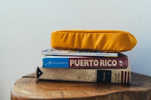 A stack of books and a magazine is topped with a bright yellow cushion. The titles visible include a guide to Puerto Rico and an adventure guidebook. The books are placed on a wooden surface with natural wood grain visible.