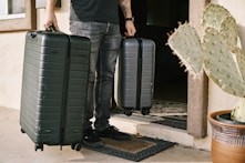 man in black denim jeans and black leather shoes standing beside black luggage bag