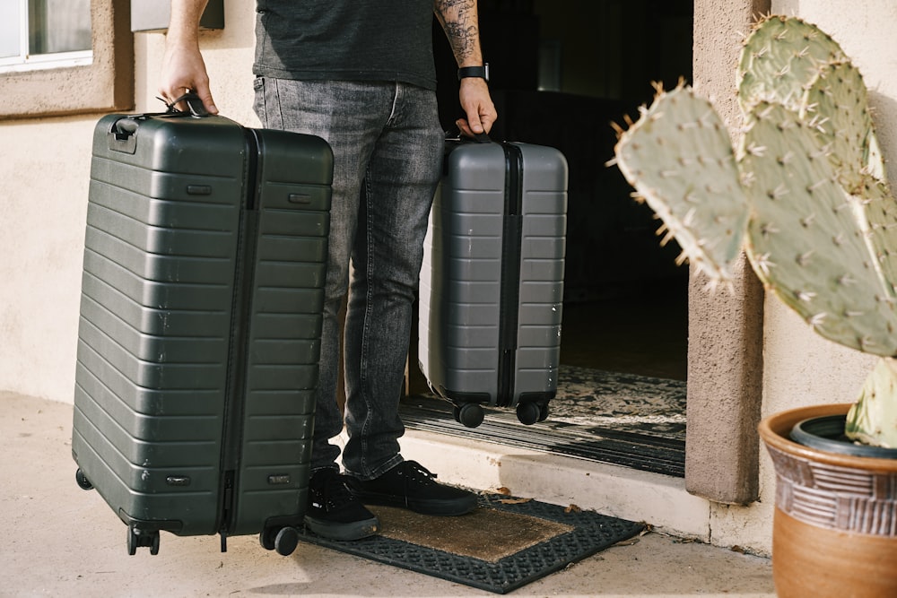 man in black denim jeans and black leather shoes standing beside black luggage bag